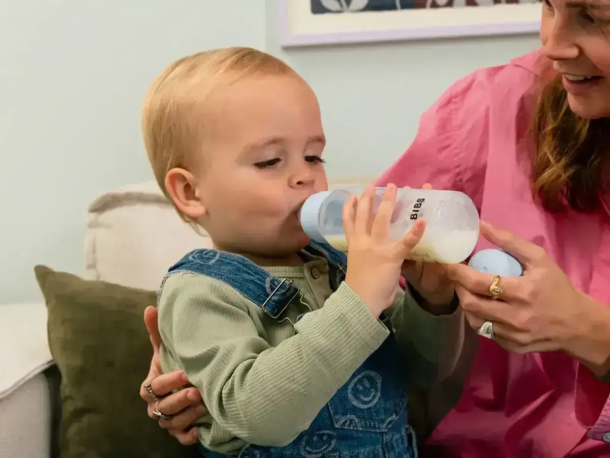 Roczne dziecko w ogrodniczkach pije mleko z butelki, którą trzyma mama.