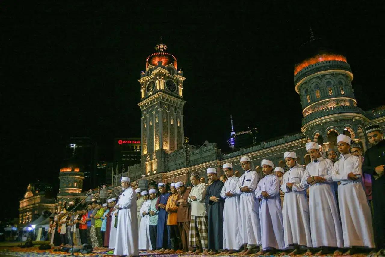 M&auml;nner und Jungen beten gemeinsam das Laylatul Qadr Gebet vor einem beleuchteten historischen Geb&auml;ude bei Nacht.