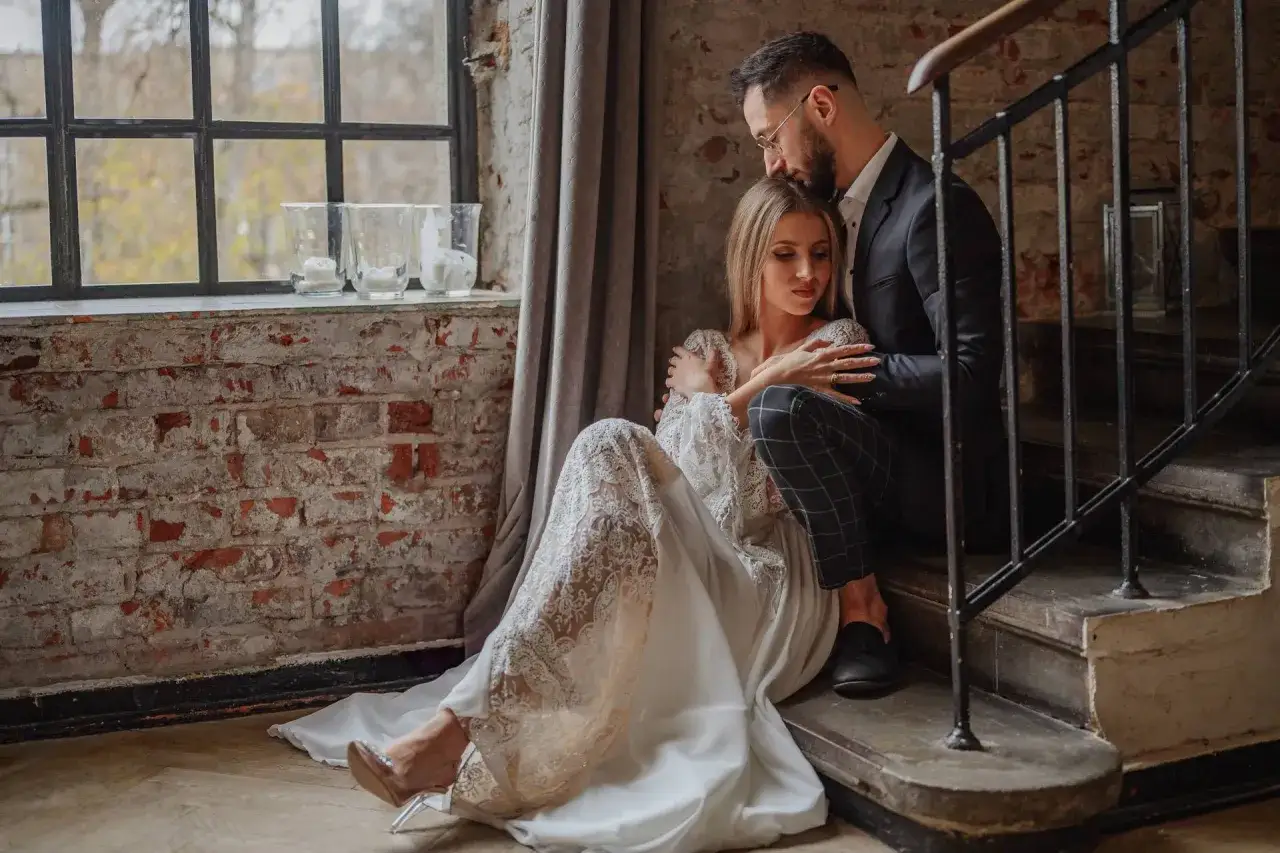Para dowiedzieć się, ile kosztuje fotograf na wesele, warto zobaczyć takie ujęcia. Para siedzi na schodach przy oknie.