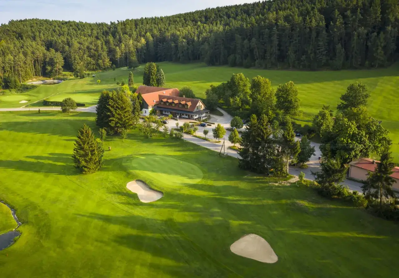 Grüne Fairways und ein Clubhaus im Schatten von Bäumen prägen den Golfclub Oberberg. Sandbunker und ein Teich sind auf dem gepflegten Rasen zu sehen.