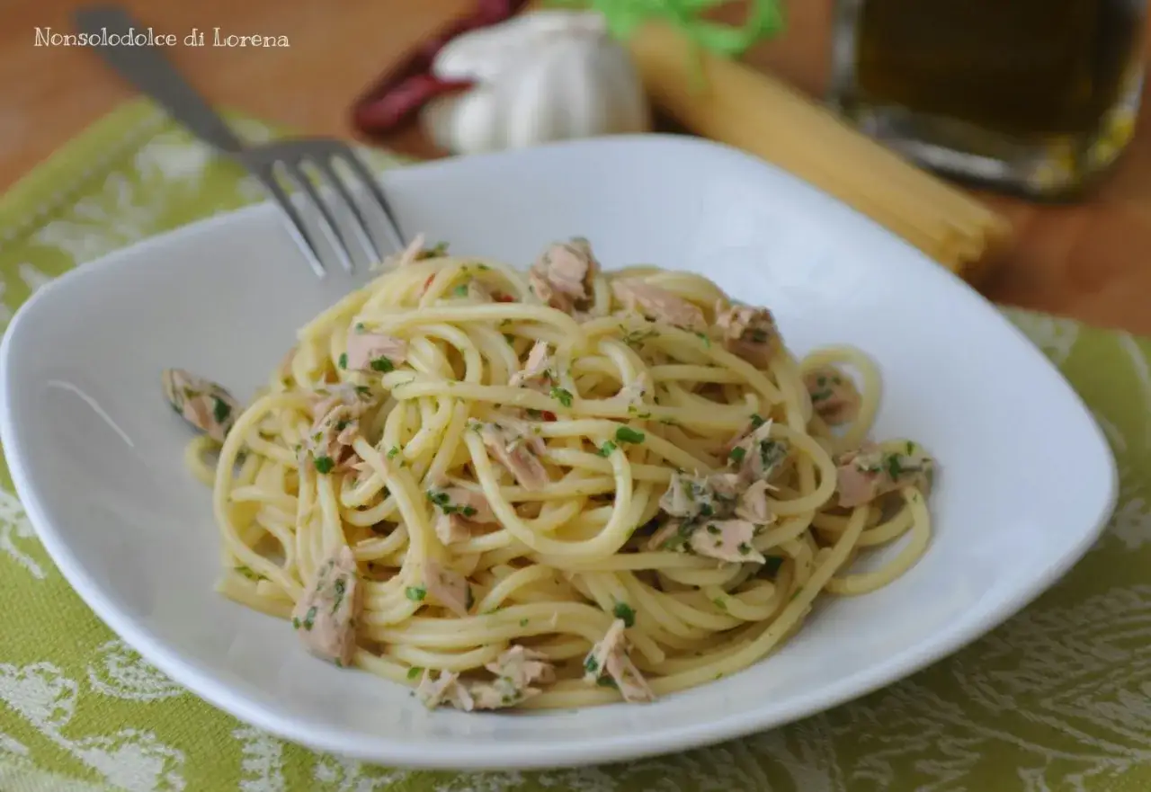 Un piatto di spaghetti con tonno e limone, conditi con prezzemolo fresco. Sullo sfondo, ingredienti per un pasto gustoso.