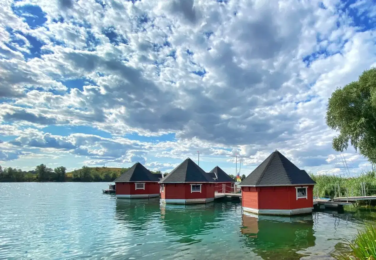 Vier rote, achteckige H&auml;uschen schwimmen auf dem Alperstedter See unter einem dramatischen Wolkenhimmel.
