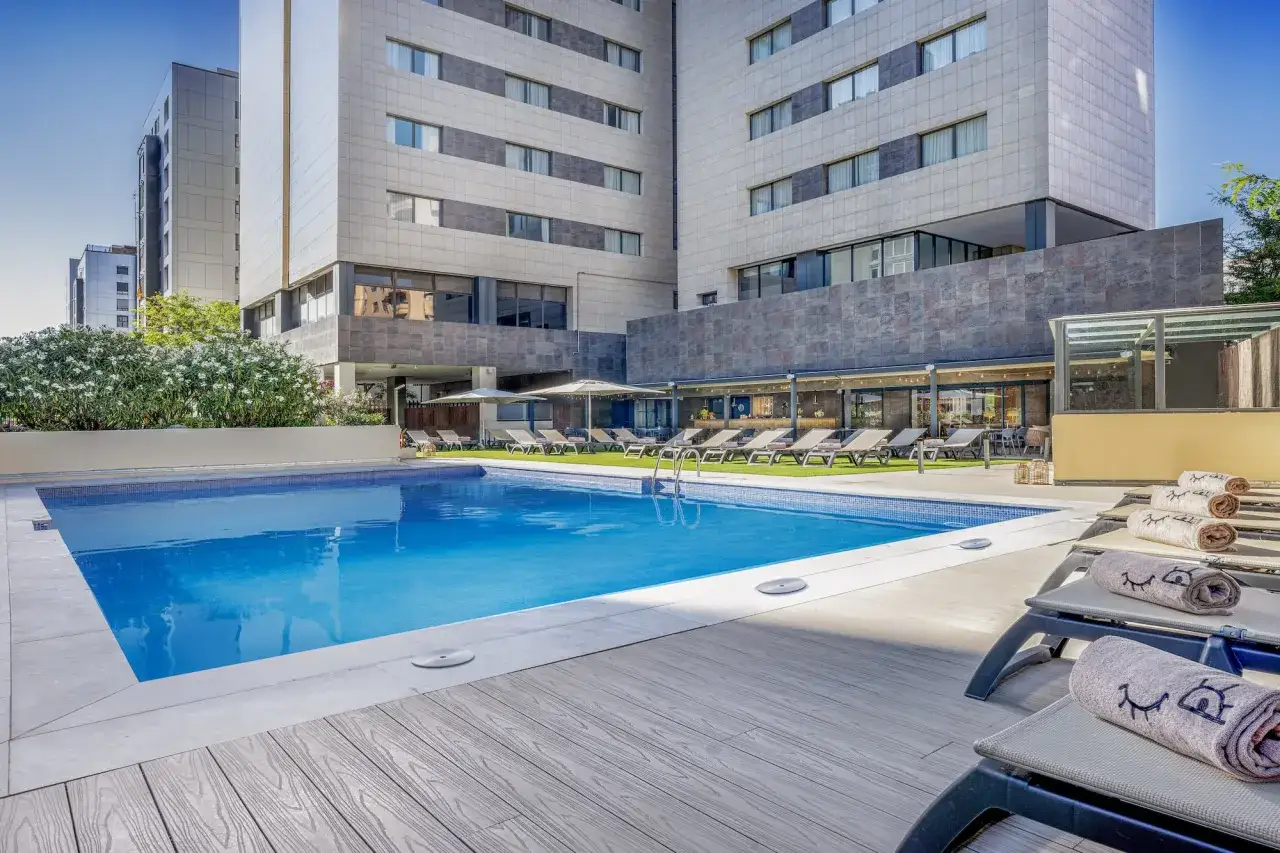 Piscina azul y tumbonas en el Meliá Valencia, rodeadas de vegetación y edificios modernos.