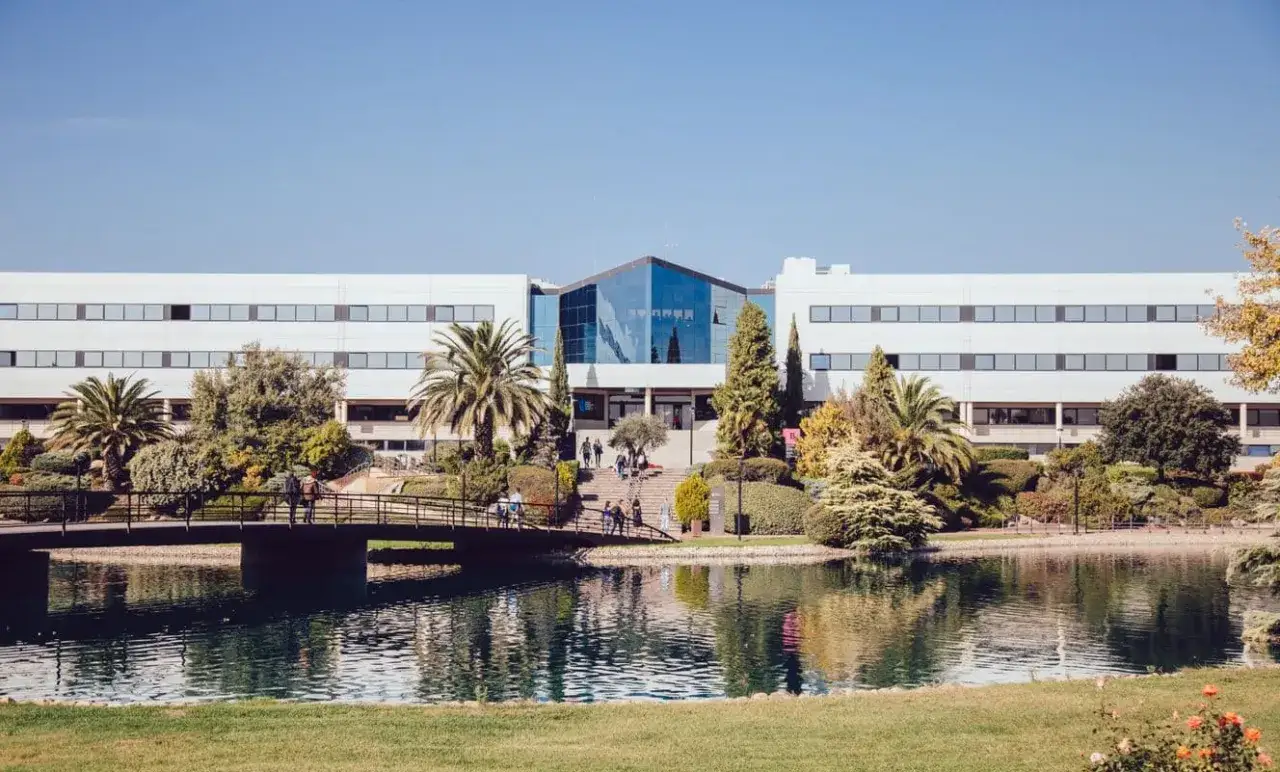 Un campus verdoyant avec un étang paisible et un pont menant à un bâtiment moderne, évoquant une **école d'élite à Madrid**.