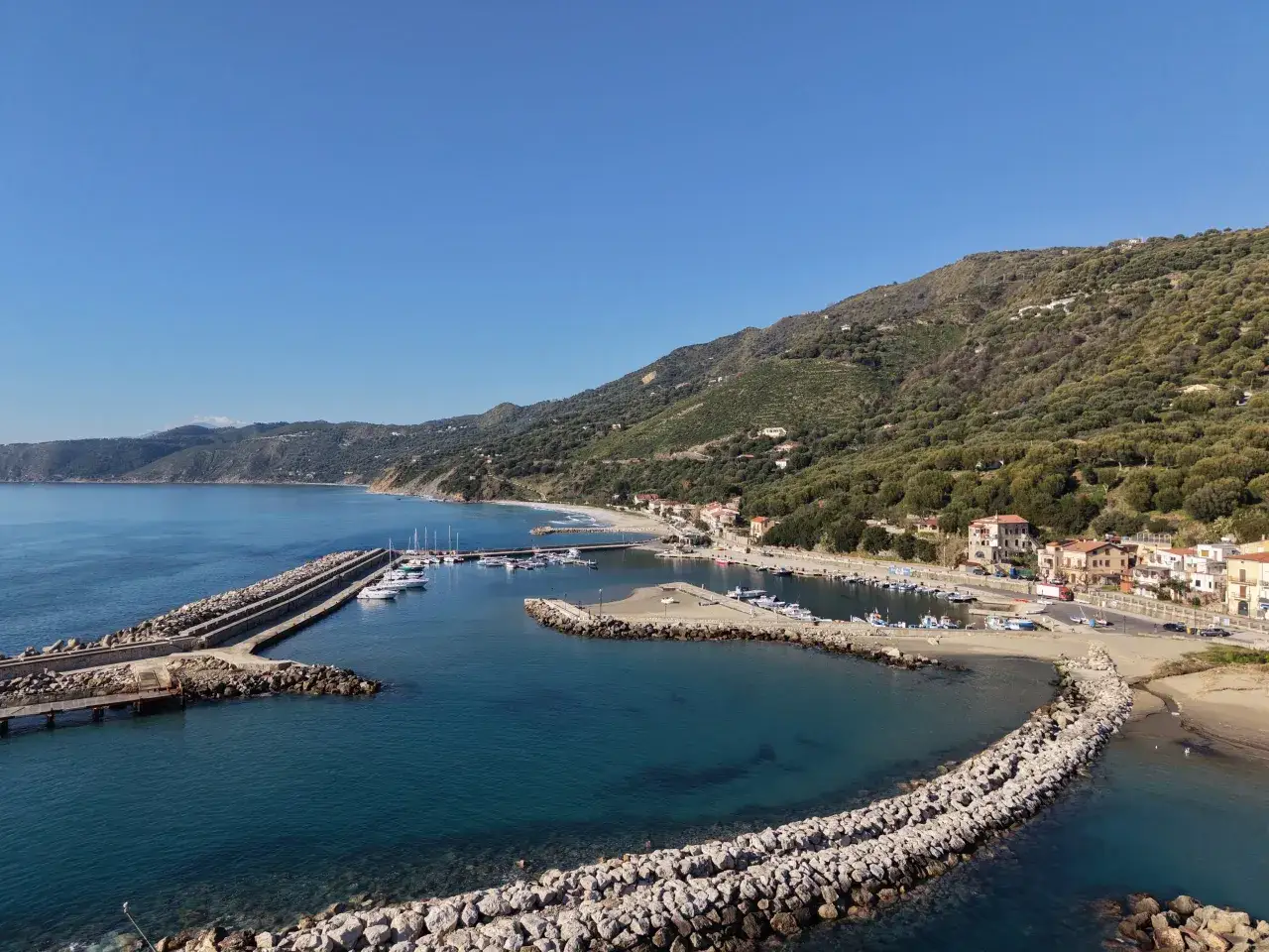 Il porto di Marina di Pisciotta, con barche ormeggiate e colline verdi sullo sfondo, sotto un cielo azzurro.