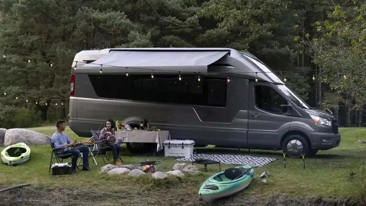Pareja disfruta de una cena al aire libre junto a una **vw california el&eacute;ctrica** en un entorno boscoso, con luces y un kayak.