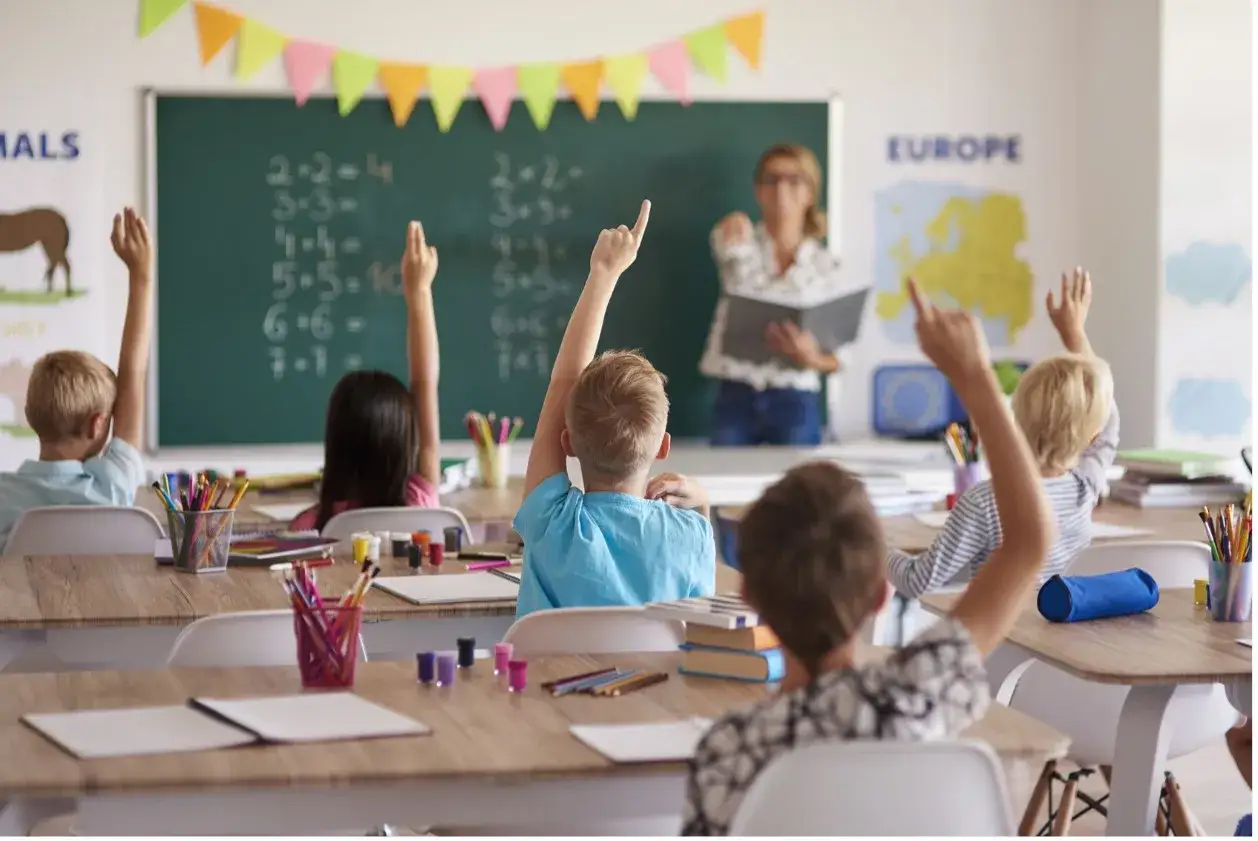 Bambini alzano la mano per rispondere, segno di come fare una lezione efficace. La maestra &egrave; in piedi davanti alla lavagna.