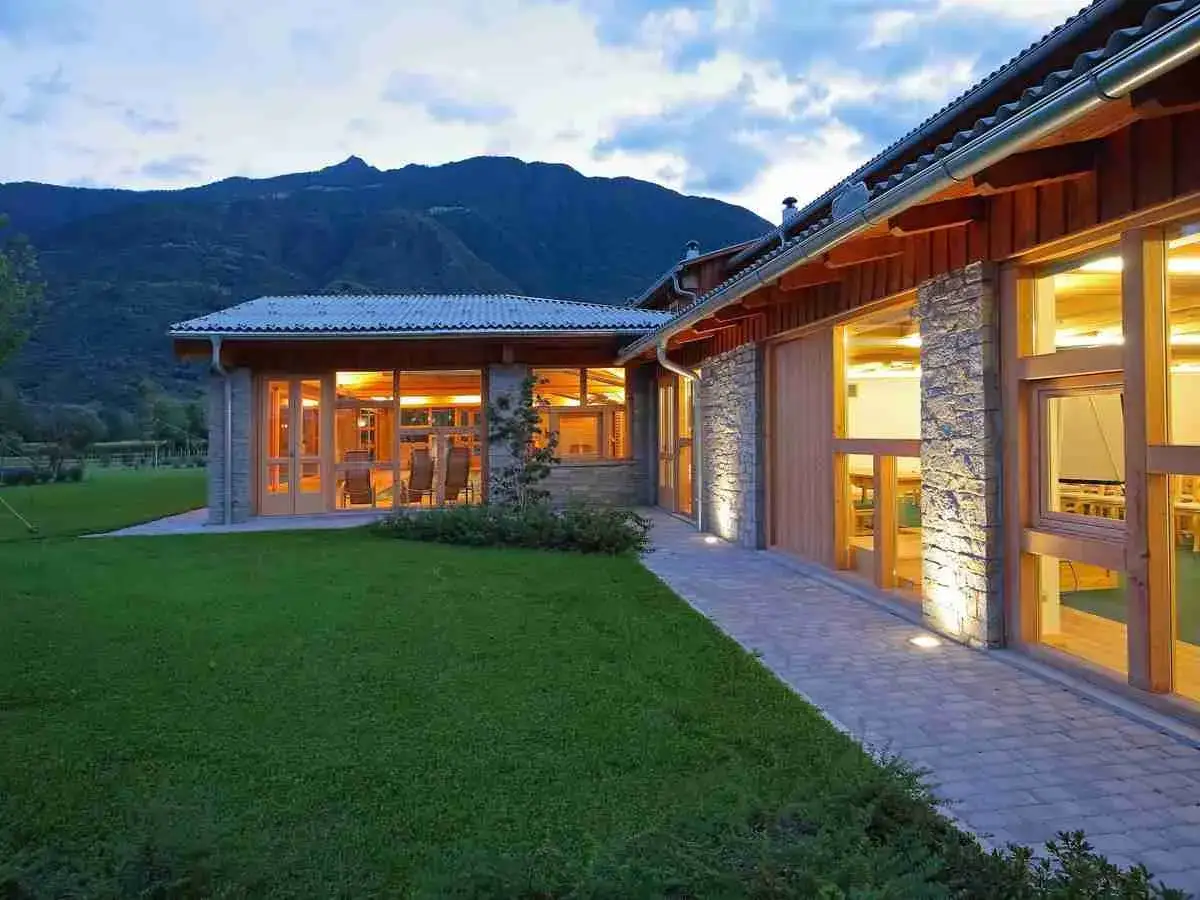 Ristorante con fattoria immerso nel verde, con ampie vetrate illuminate che invitano all'interno. Montagne sullo sfondo.