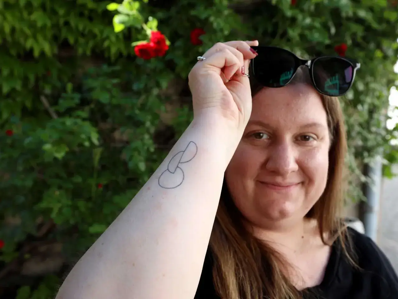Frau mit einem Organ&shy;spende-Tattoo auf dem Arm, l&auml;chelnd vor einer gr&uuml;nen Hecke mit roten Rosen.