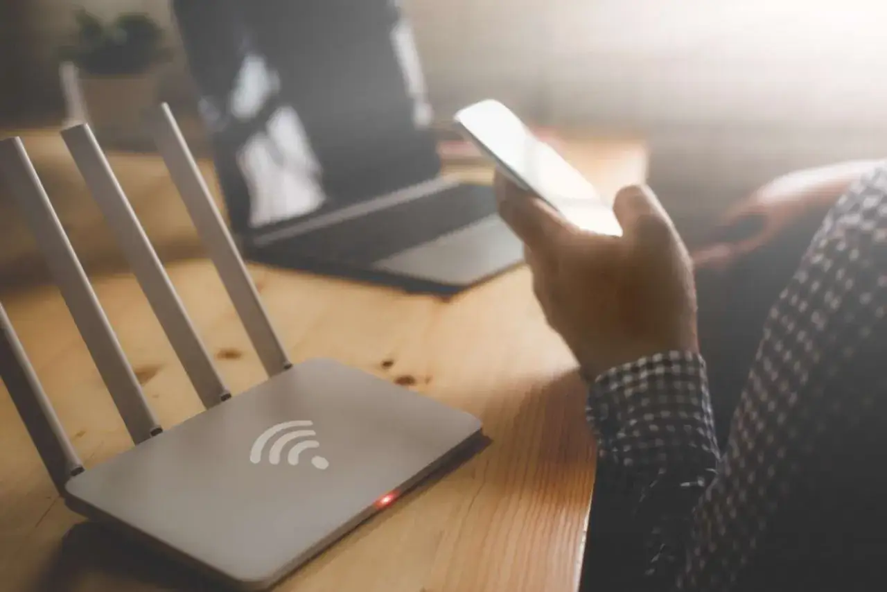 Router z ikoną wlan i świecącą diodą, obok smartfon w dłoni i laptop.