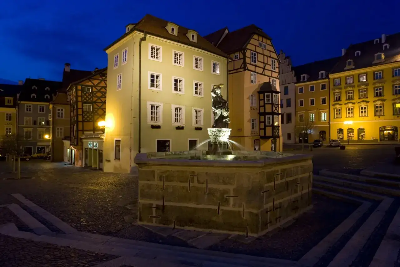 Kamienny rynek w **Cheb** nocą, z podświetlaną fontanną i zabytkowymi kamienicami.