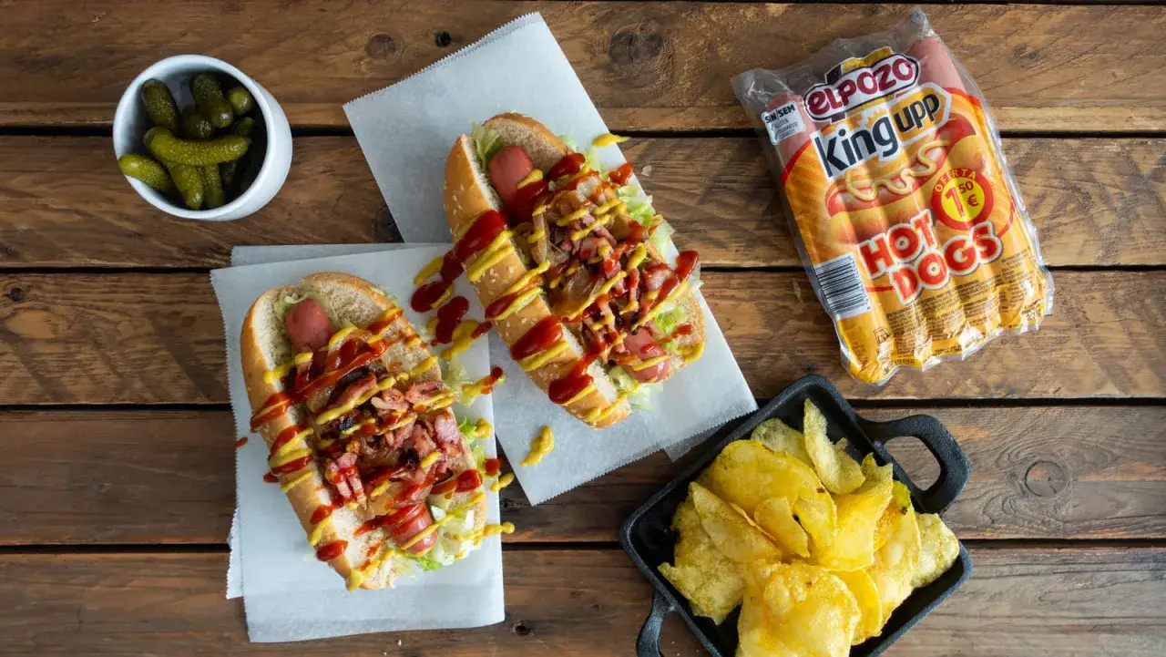 Dos perritos calientes con lechuga, bacon, cebolla, ketchup y mostaza. Acompañados de pepinillos y patatas fritas.