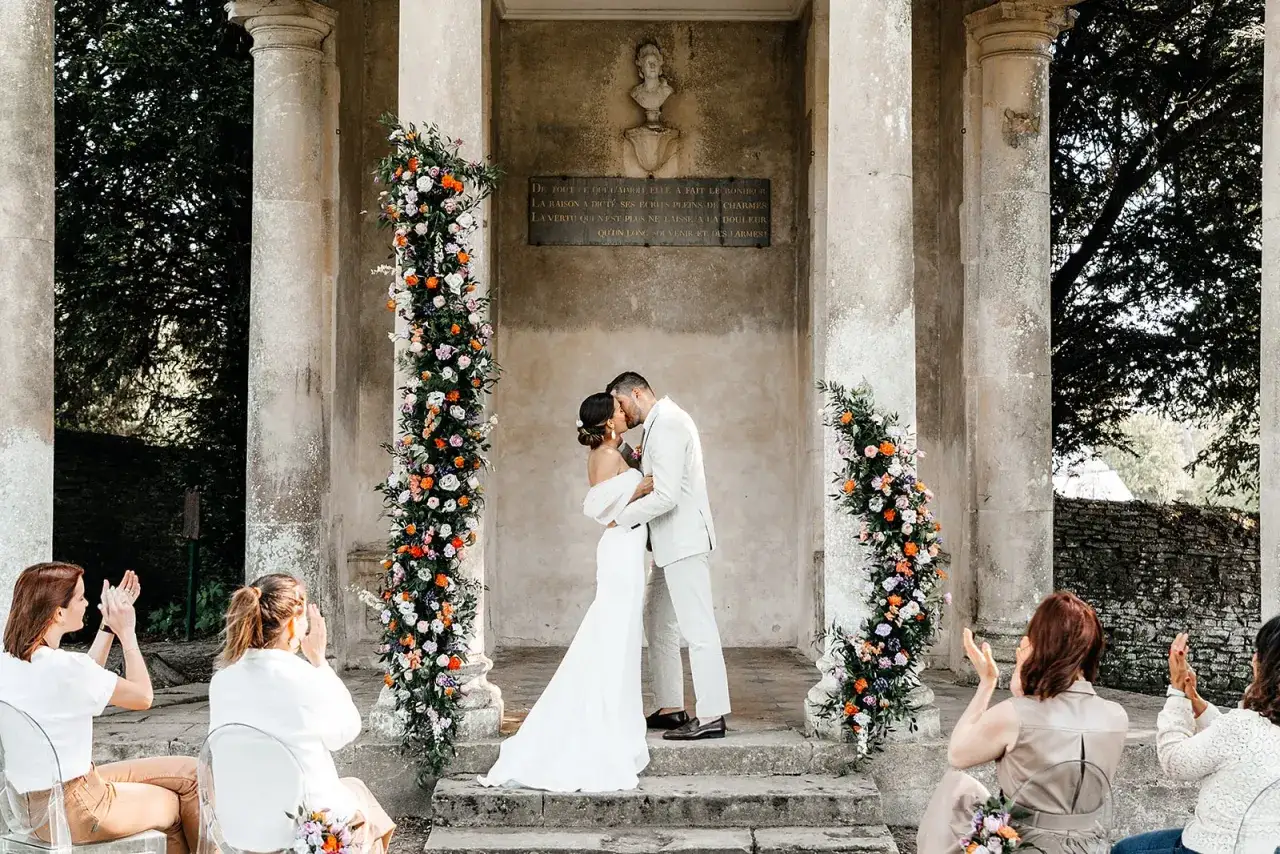 Un couple s'embrasse lors d'un mariage Bridgerton, entour&eacute; de fleurs color&eacute;es. Des invit&eacute;s applaudissent, cr&eacute;ant une sc&egrave;ne romantique et &eacute;l&eacute;gante.