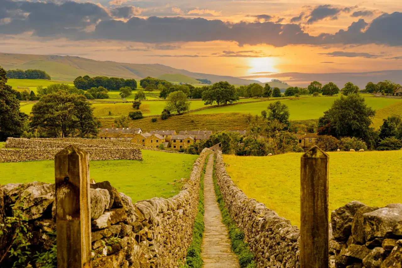 A stone wall path leads through green fields towards a village nestled in the hills, bathed in the warm glow of a sunset. This is the Dales Way.