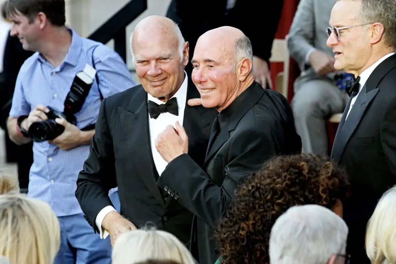 Two men in tuxedos smile, perhaps discussing their favorite pieces from the David Geffen art collection. A photographer is in the background.