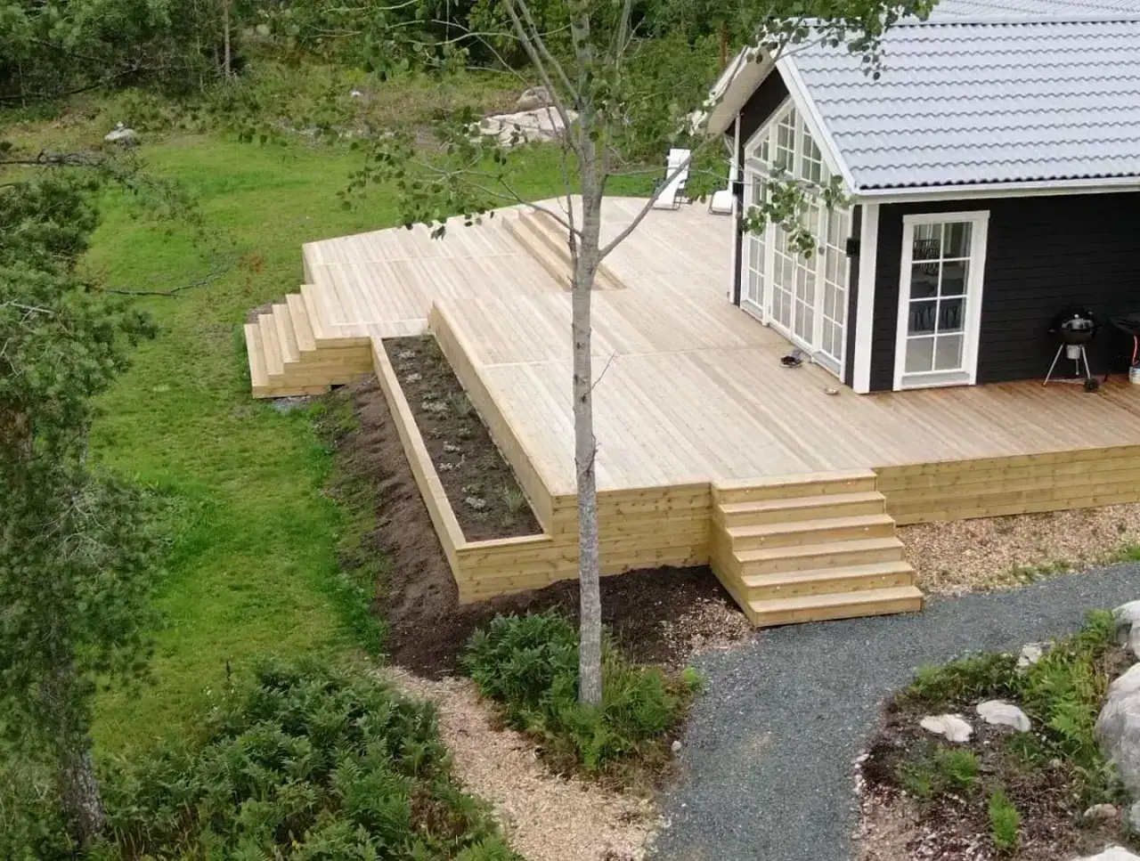 Nybyggd altan i tr&auml; som ramar in ett svart hus. Perfekt f&ouml;r att bygga uteplats och njuta av naturen.