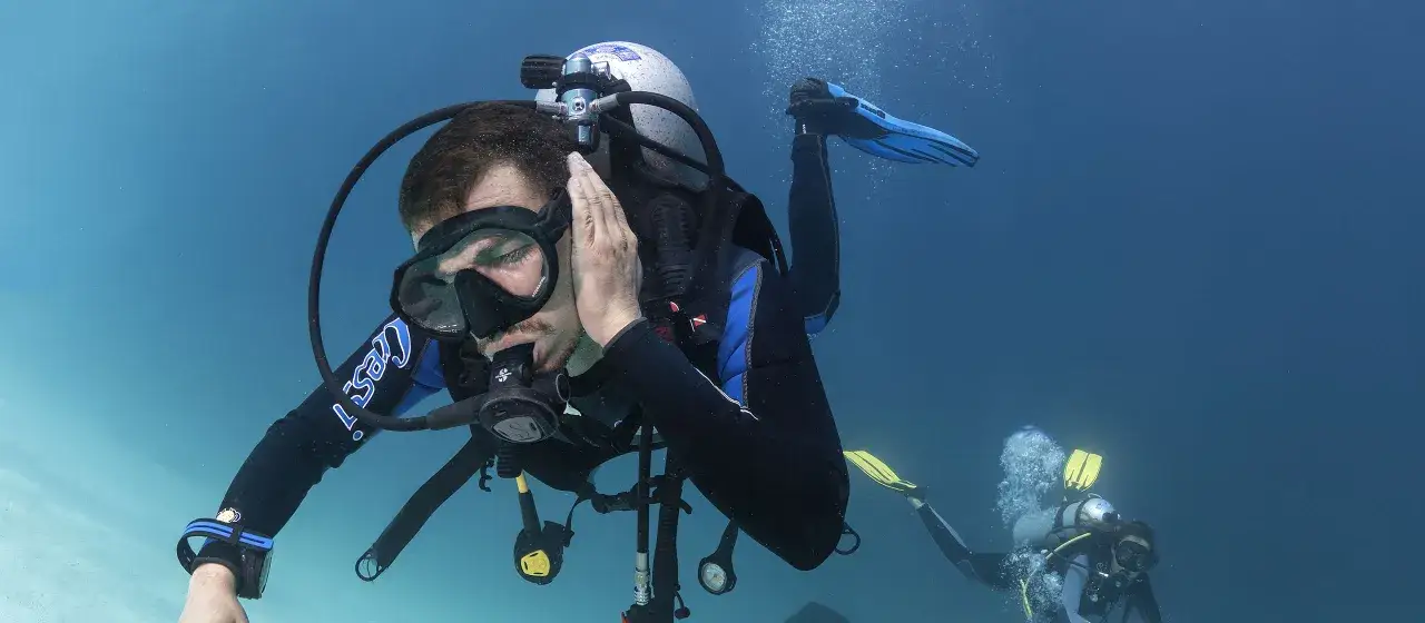 Nurkowie pod wodą, jeden z nich dotyka ucha, być może doświadczając barotrauma. Co to jest?