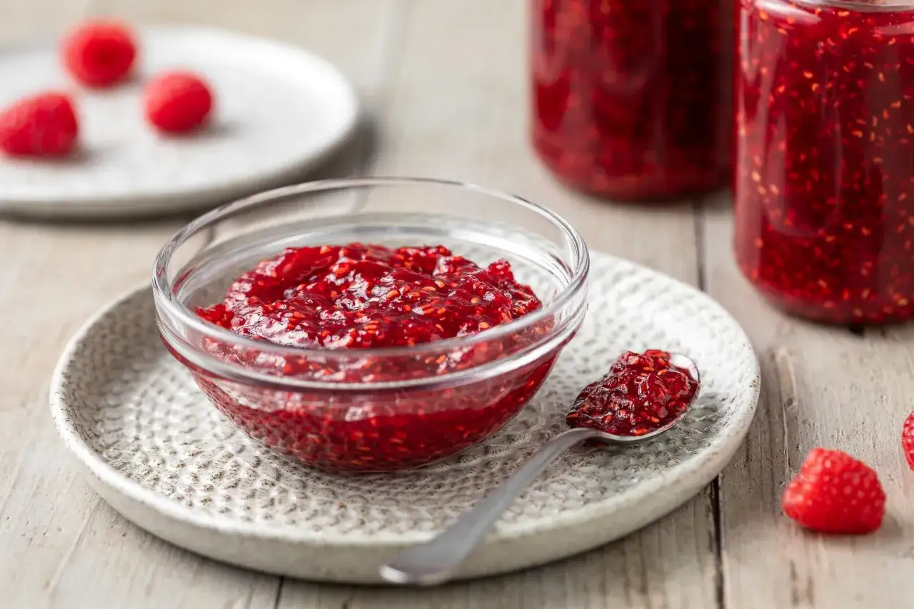 Ciotolina di marmellata di lamponi fatta in casa, con un cucchiaino pieno e lamponi freschi sullo sfondo.