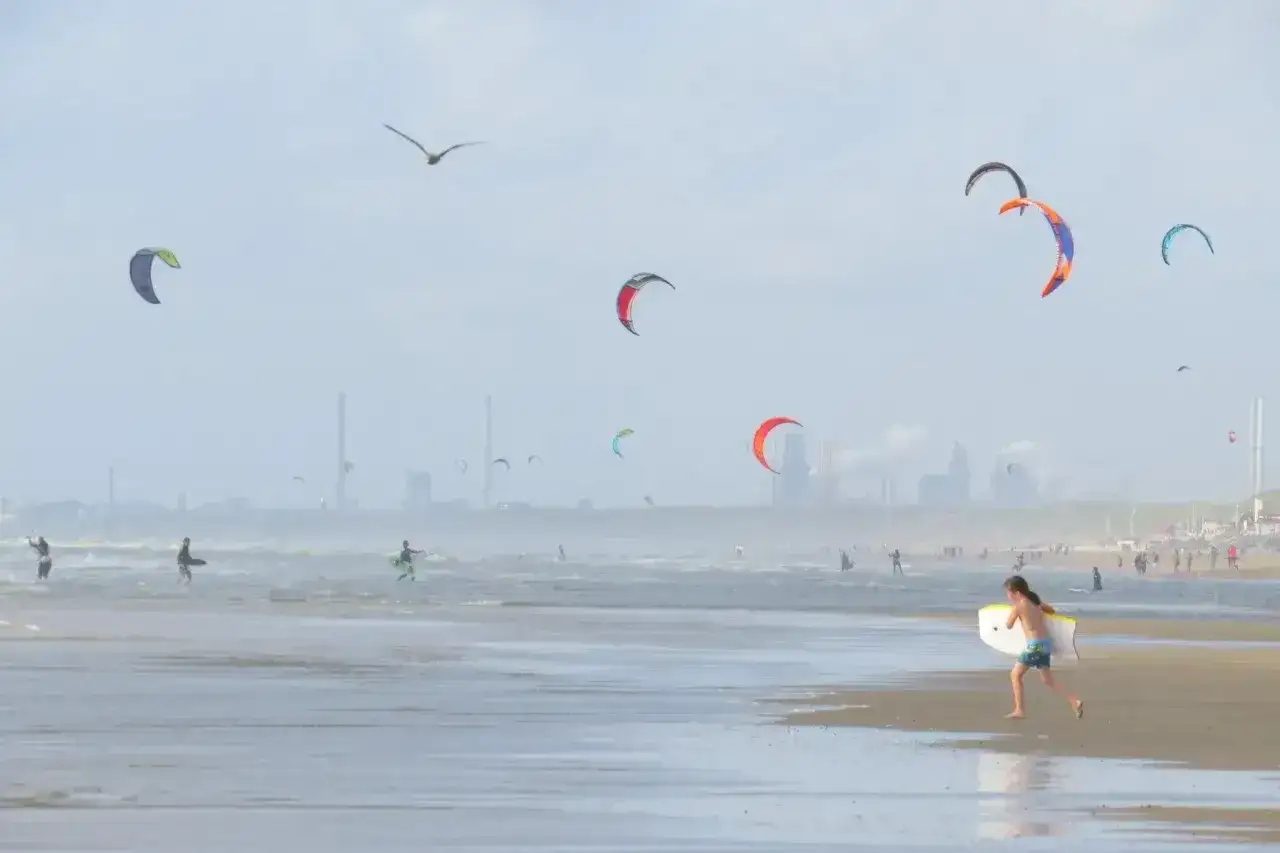 Dziecko z deską biegnie po plaży w Holandii, gdzie na morzu trwa kitesurfing. W tle widać przemysłowe budynki i mewę.