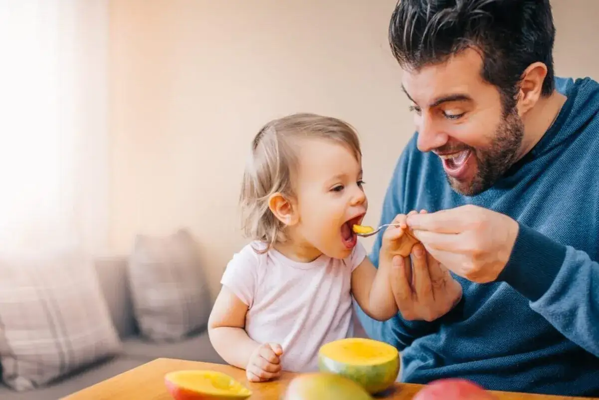 Padre sonriente alimenta a su bebe de 8 semanas con mango.