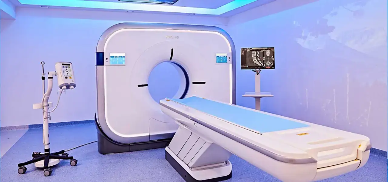 Nowoczesny aparat do tomografii głowy w jasnym pomieszczeniu. Obok leżanka pacjenta i monitor z obrazami diagnostycznymi.