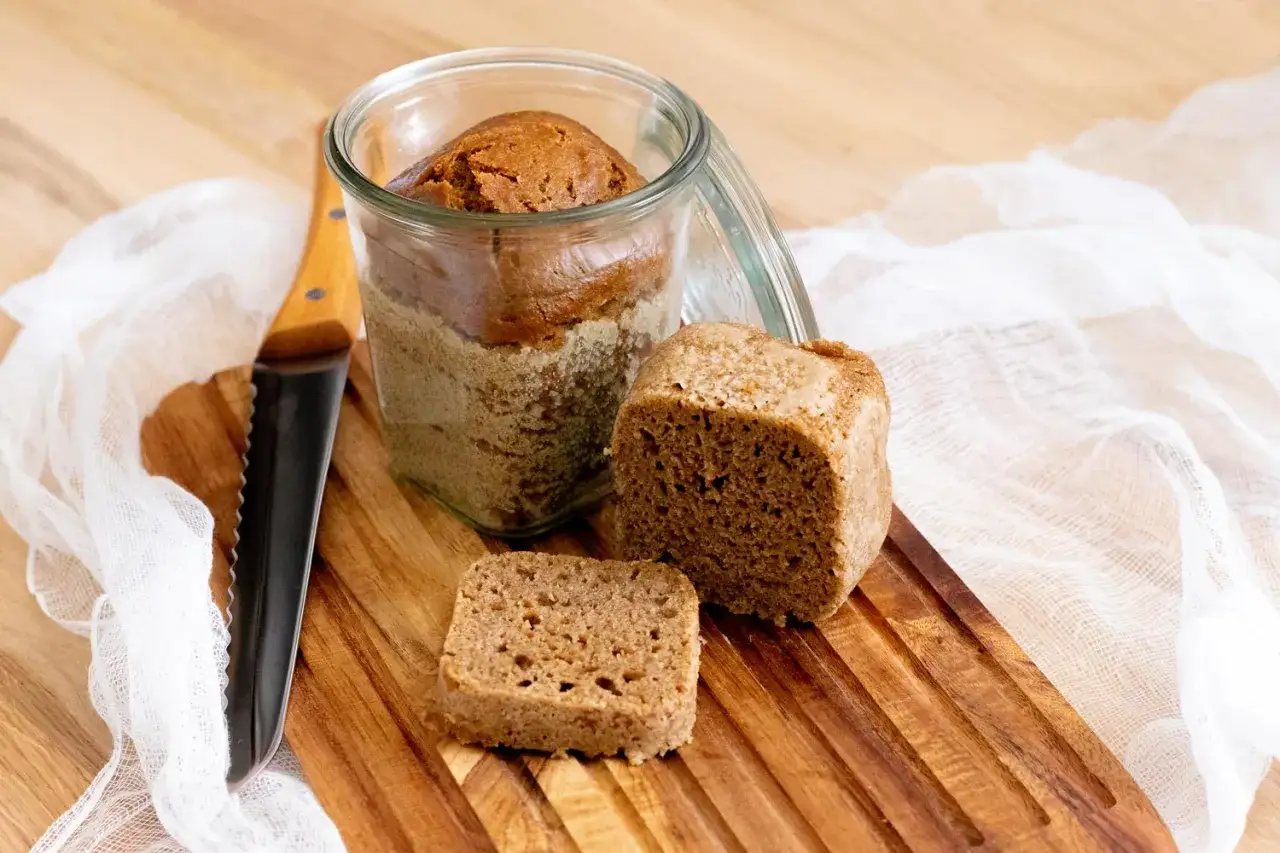 Brot im Glas Rezept: Ein Laib Brot ist im Glas, daneben liegen ein Stück und ein Krümel auf einem Holzbrett. Ein Messer liegt daneben.