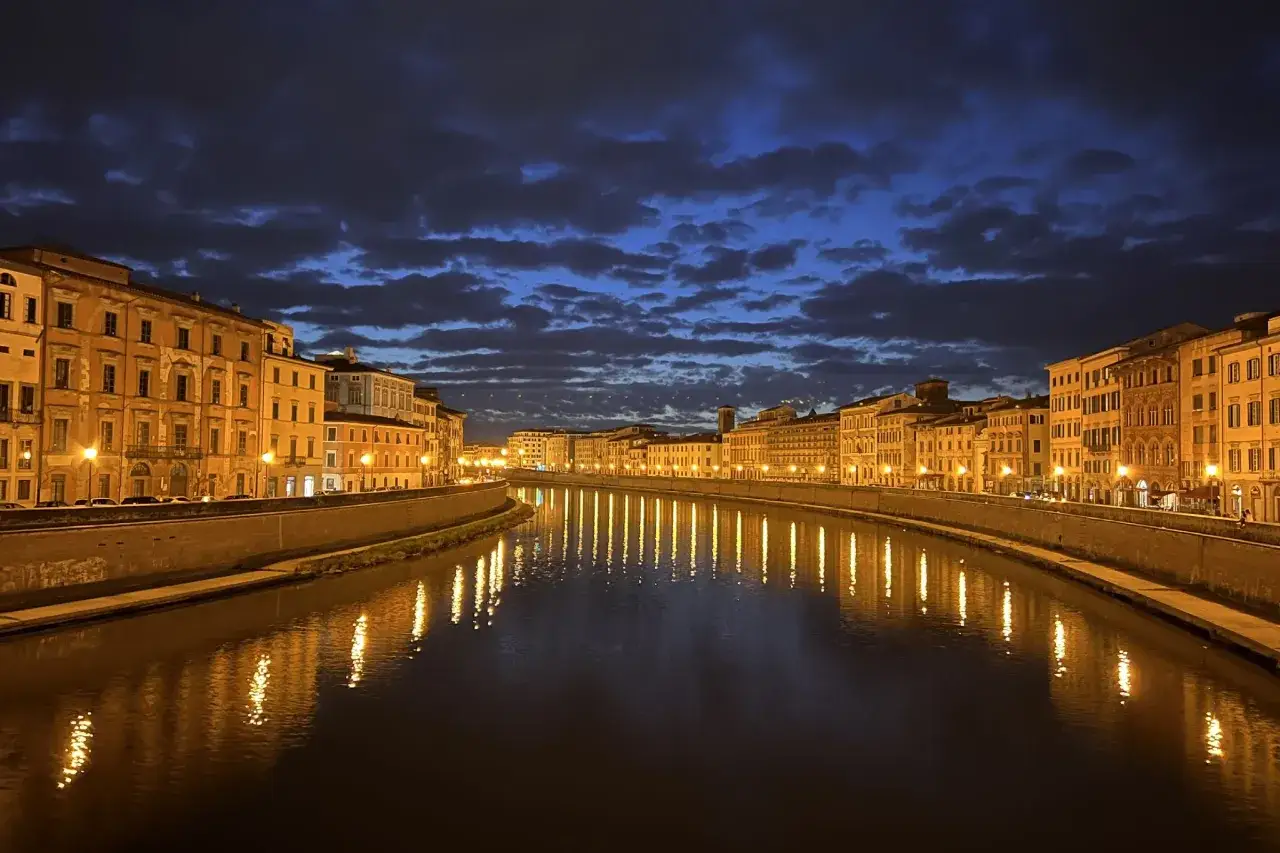 Edifici illuminati lungo il fiume Arno a Pisa, con riflessi dorati sull'acqua scura sotto un cielo notturno nuvoloso. Un'atmosfera magica per scoprire cosa ver en Pisa.