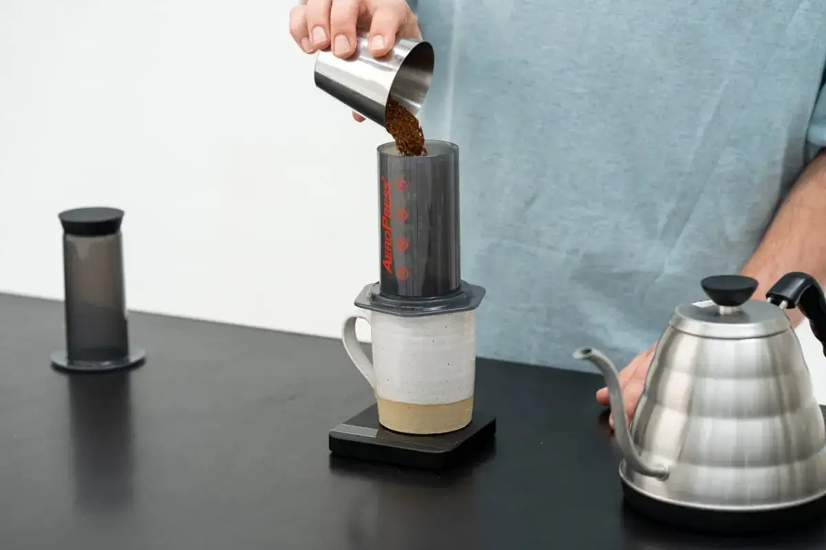 Kaffeezubereitung mit AeroPress: Gemahlener Kaffee wird aus einem Metallbecher in den AeroPress-Zylinder gegossen.
