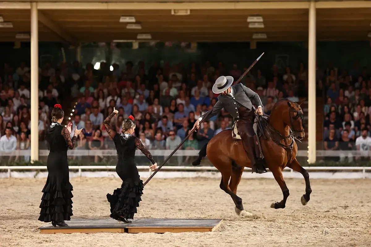 Spectacle de doma vaquera : un cavalier en costume traditionnel interagit avec deux danseuses de flamenco sur une piste de sable, devant un public nombreux.