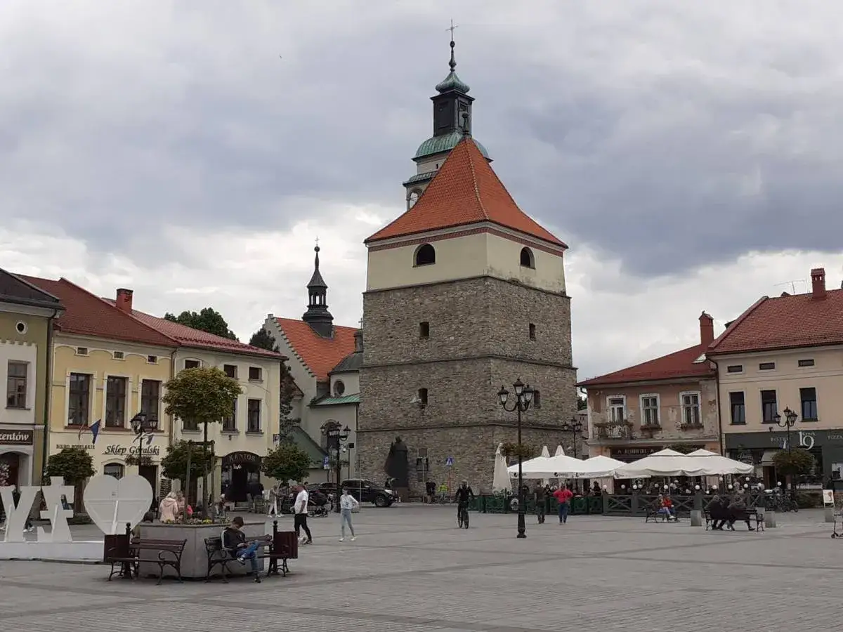 Żywiec rynek z wieżą ratuszową i kamienicami. Ludzie spacerują, siedzą na ławkach, a kawiarnie zapraszają pod białe parasole.