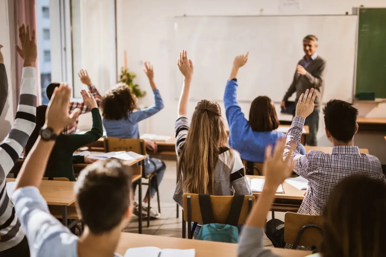 Uczniowie z podniesionymi rękami, gotowi do odpowiedzi. Nauczyciel czeka na pytania. To świetny wybór obrazka po angielsku zwroty, pokazujący zaangażowanie w lekcję.