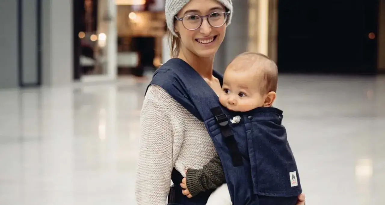 Uśmiechnięta mama w czapce i okularach nosi dziecko w granatowym nosidełku. Idealne rozwiązanie, gdy zastanawiasz się, jakie nosidełko wybrać.