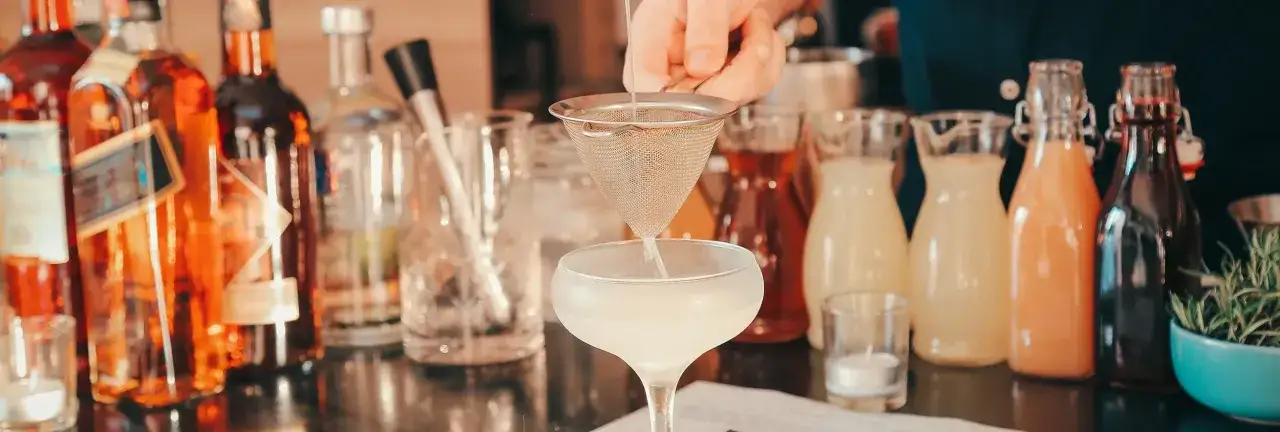 Barman przygotowuje drinka, filtrując płyn do kieliszka. Obok stoją butelki z alkoholami i syropami, idealne na wesele. Ile w&oacute;dki na wesele? To zależy od gości!