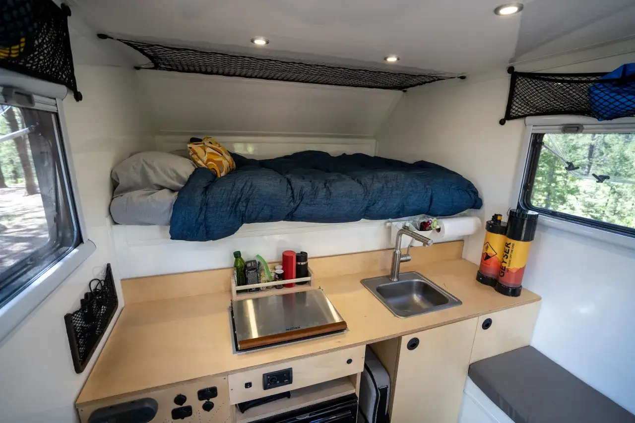 Int&eacute;rieur d'une cellule pick-up am&eacute;nag&eacute;e : lit douillet, coin cuisine avec &eacute;vier et r&eacute;chaud, et rangements nets. Parfait pour l'aventure !