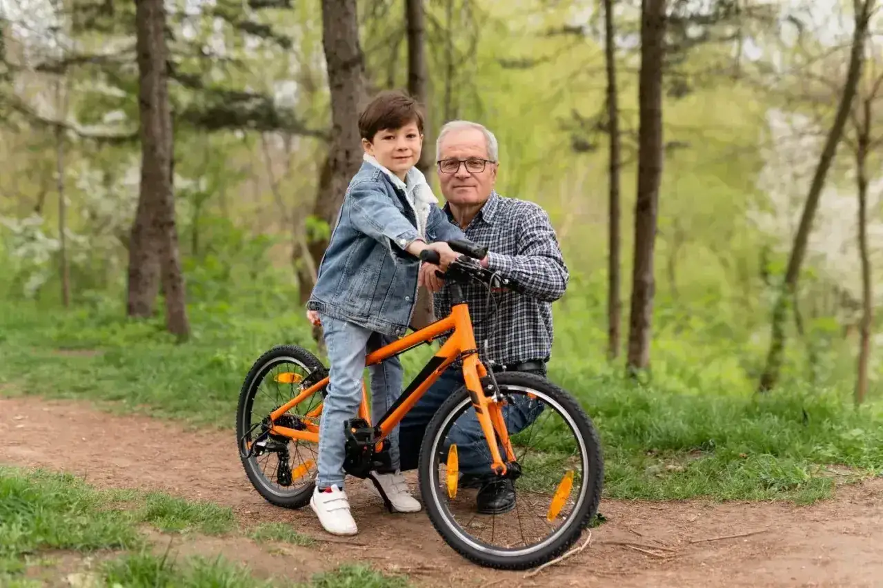 Dziadek pomaga wnukowi wybrać rower. Chłopiec stoi przy pomarańczowym rowerze, gotowy do jazdy.