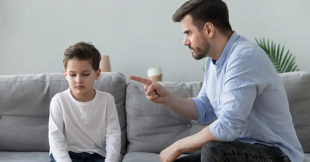 Ojciec strofuje syna, który siedzi ze spuszczoną głową. Trudne chwile w rodzicielstwie.