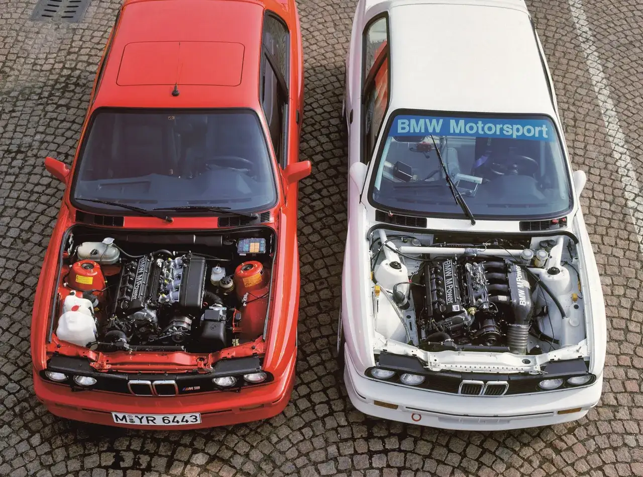 Dwa klasyczne BMW z otwartymi maskami, prezentujące moc silników BMW M Power. Czerwony i biały model stoją obok siebie na brukowanej nawierzchni.