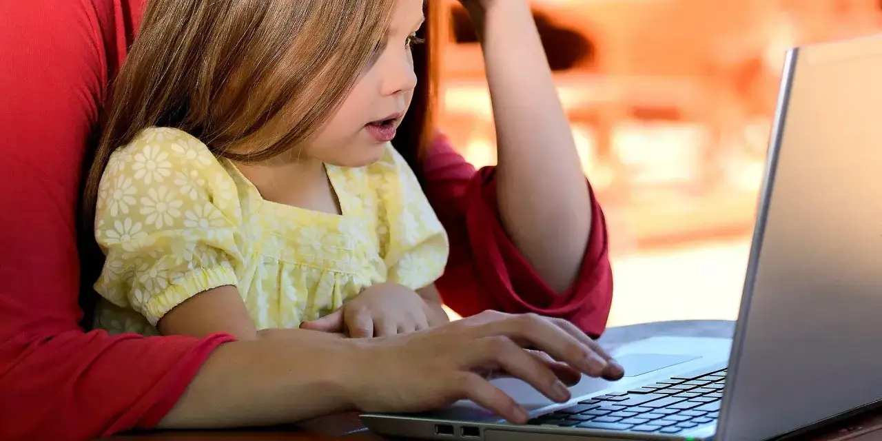 Dziecko z zaciekawieniem patrzy na ekran laptopa, podczas gdy rodzic korzysta z aplikacji do kontroli rodzicielskiej, dbając o bezpieczeństwo online.