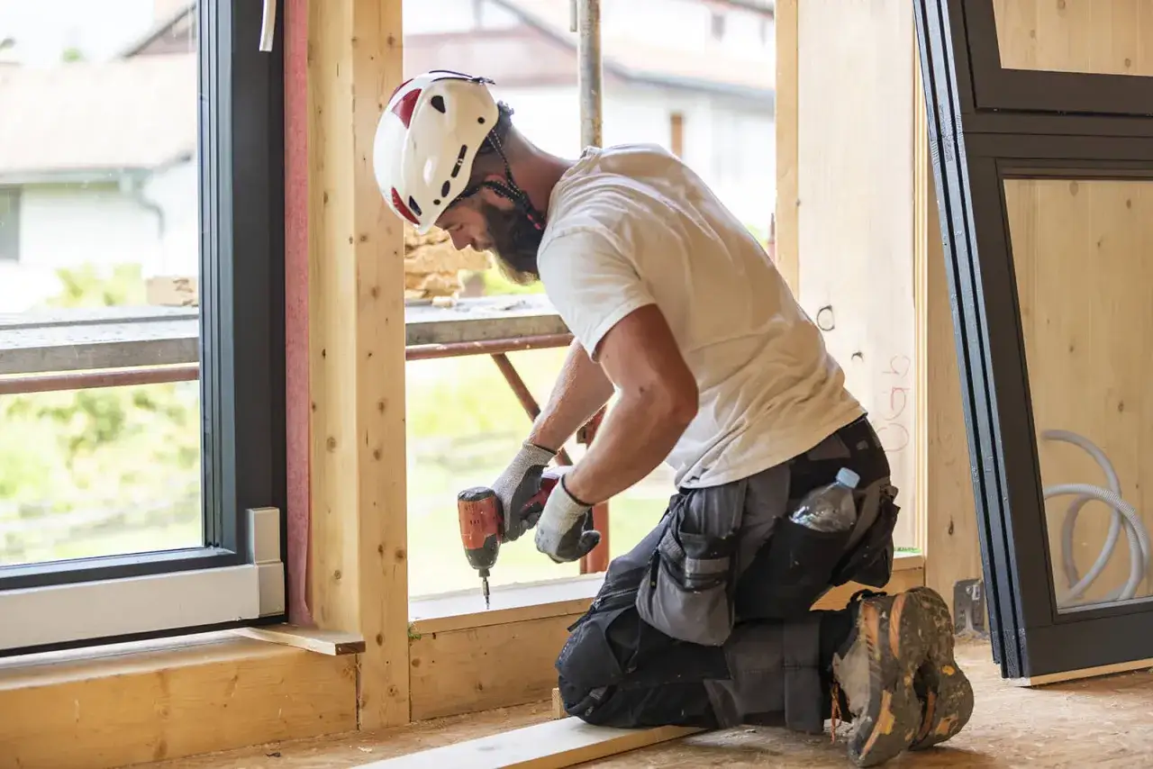 Un ouvrier en casque blanc utilise une visseuse pour changer une fenêtre. Il est agenouillé dans une maison en construction.