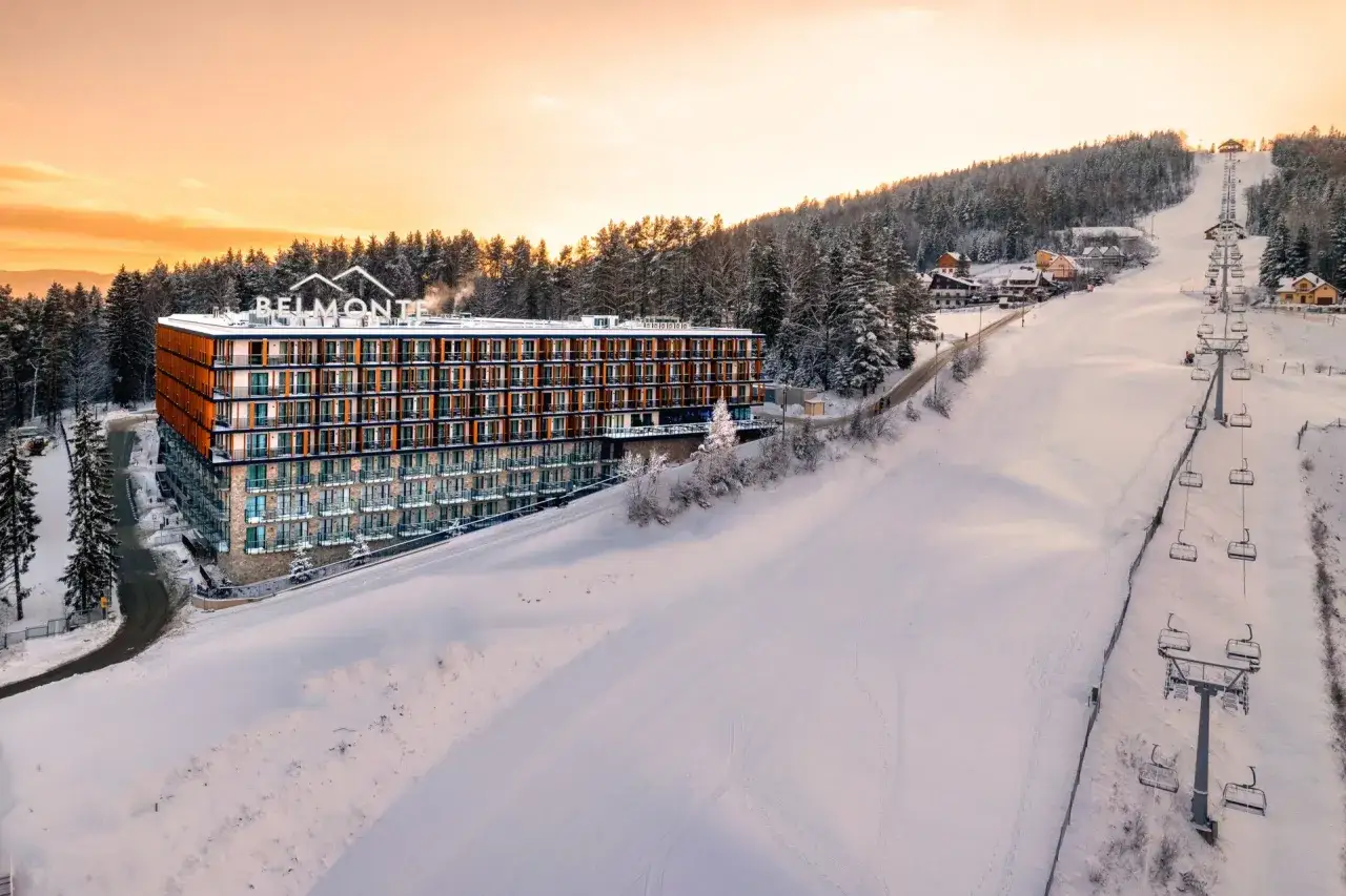 Nowoczesny hotel Belmonte przy stoku narciarskim w Polsce, otoczony ośnieżonym lasem, z widoczną kolejką krzesełkową.
