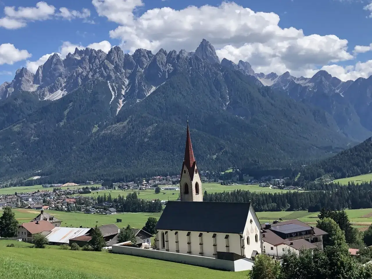 Chiesa con campanile rosso in primo piano, sullo sfondo le montagne maestose e i **paesi pi&ugrave; belli della Val Pusteria**.