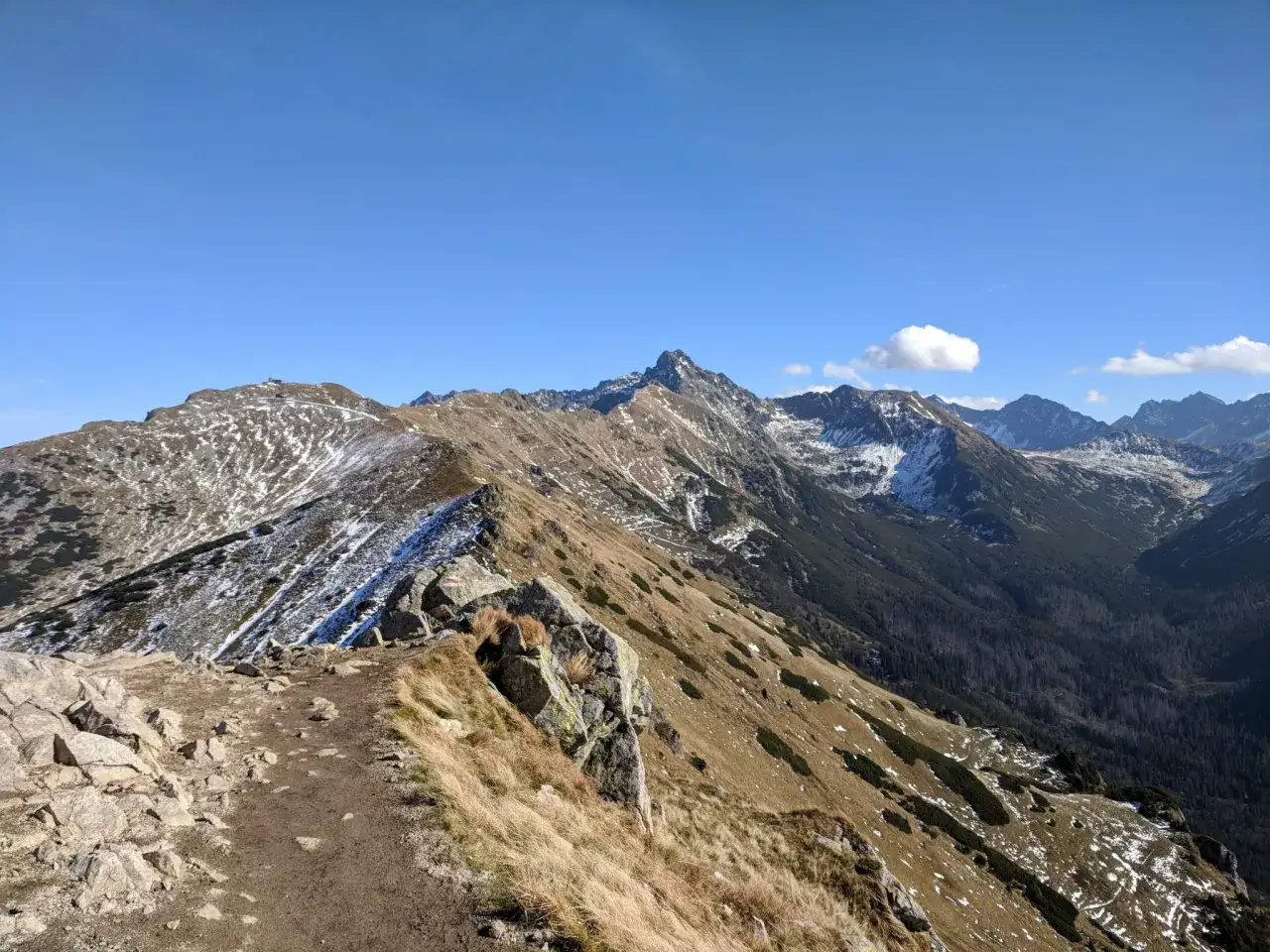 Widok na Tatry z grani, gdzie widać ślady śniegu na zboczach. W oddali majaczy Giewont, a po lewej stronie Kasprowy Wierch.