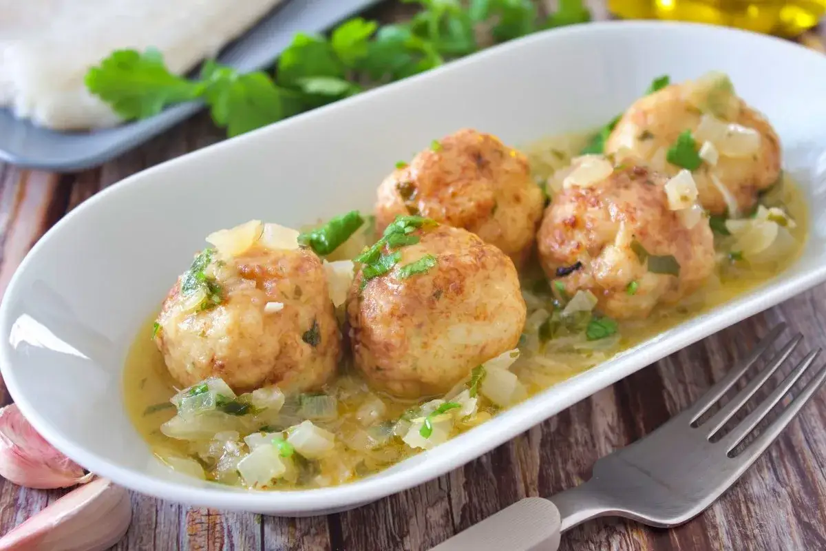 Albondigas de merluza caseras en una salsa cremosa con cebolla y perejil, servidas en plato blanco.