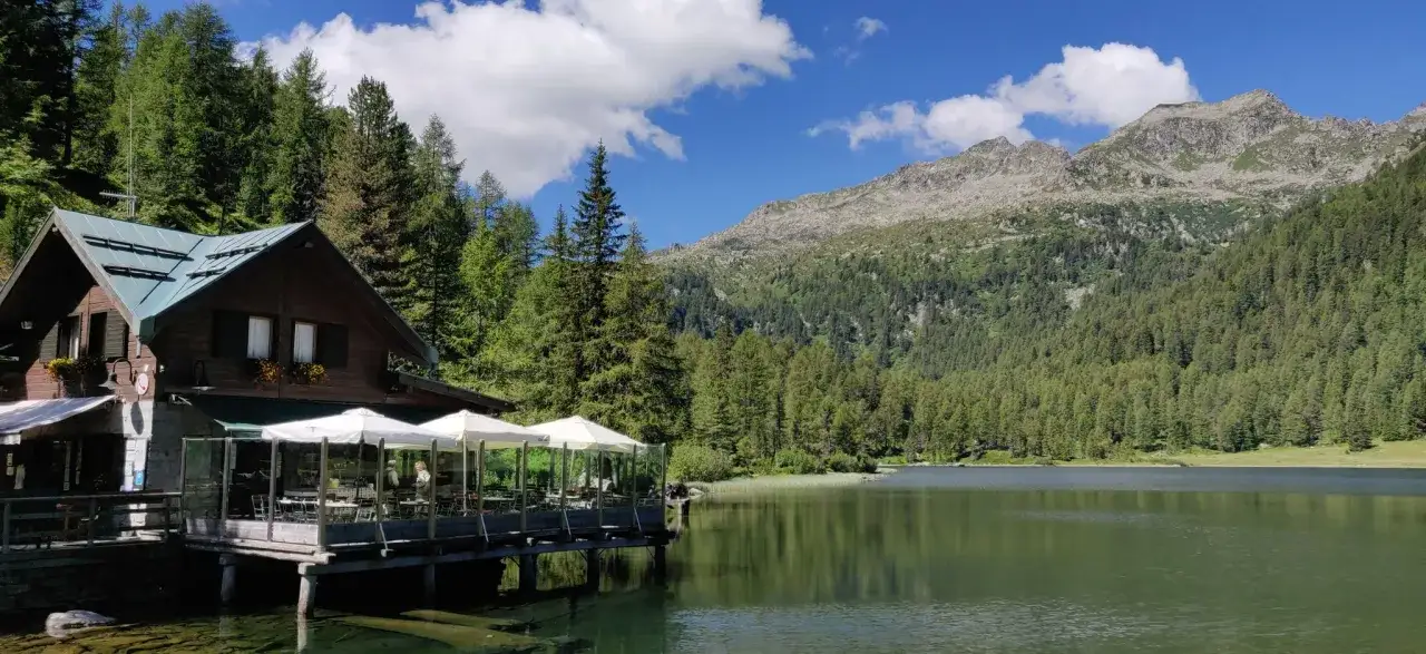 Rifugio in legno con tavoli all'aperto su un lago alpino, circondato da foreste e montagne. Un luogo incantevole per scoprire cosa vedere a Pellizzano.