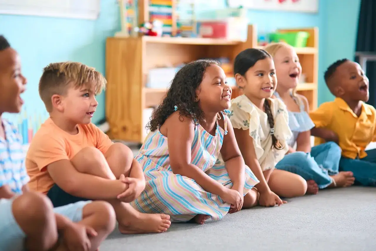 Bambini seduti in cerchio, felici e attenti, in un'aula colorata. Un momento di gioco e apprendimento condiviso.