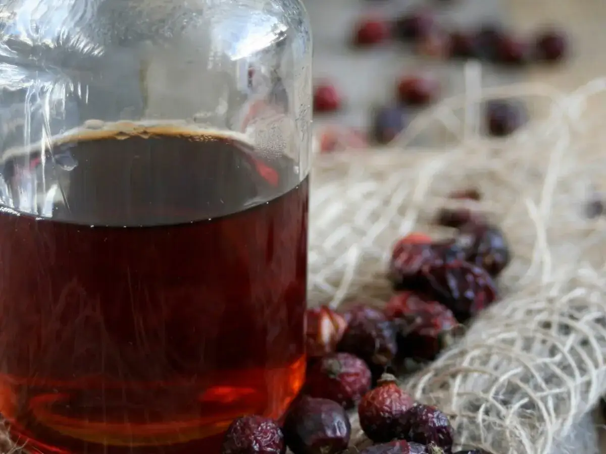 Syrop z dzikiej róży przepis. Ciemny, aromatyczny syrop w szklanej butelce, obok suszone owoce dzikiej róży na jutowym sznurku.