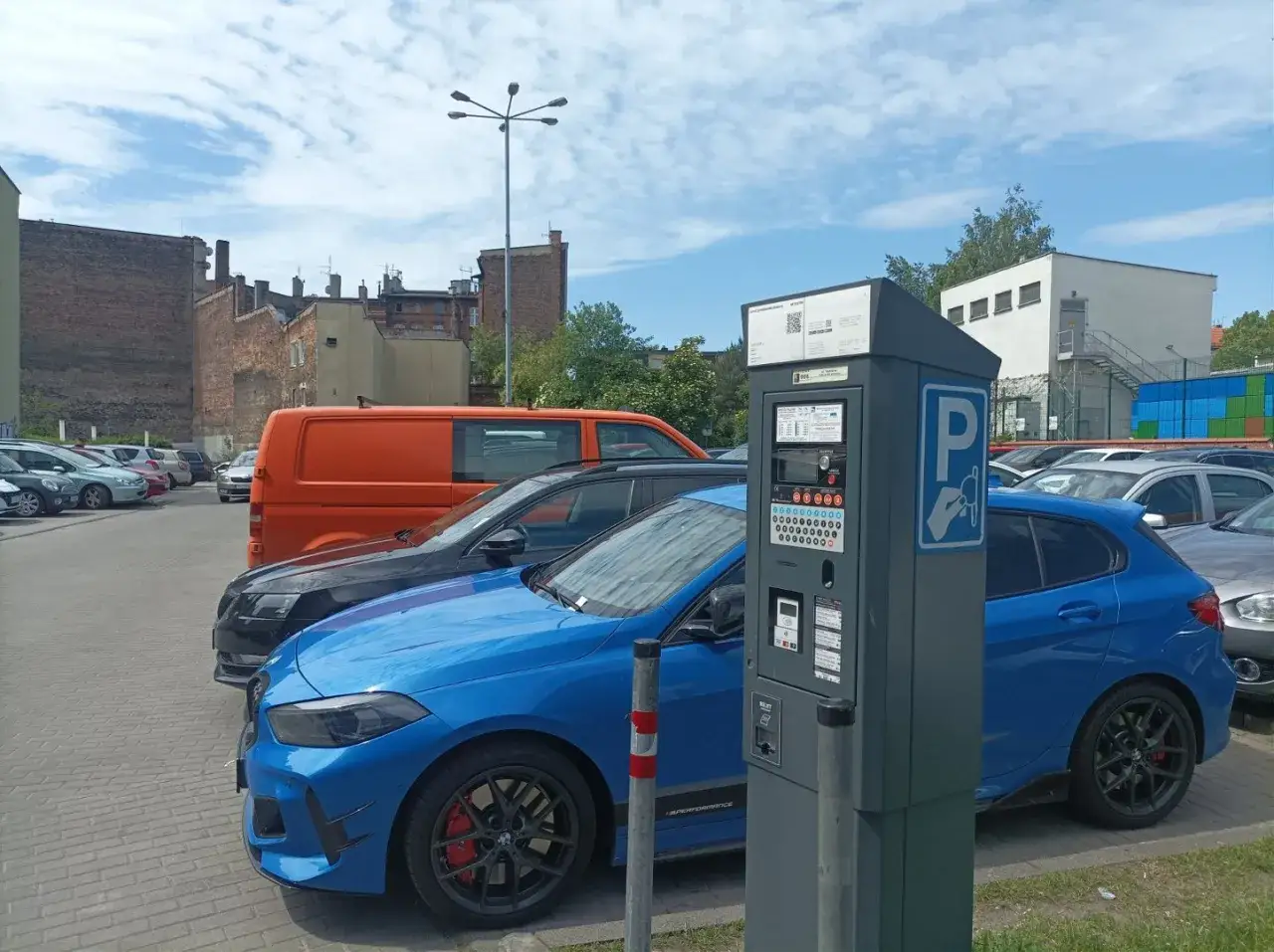 Parking przy Teatrze Rozrywki w Chorzowie. Niebieski i czarny samochód, pomarańczowy bus, parkometr i budynki w tle.