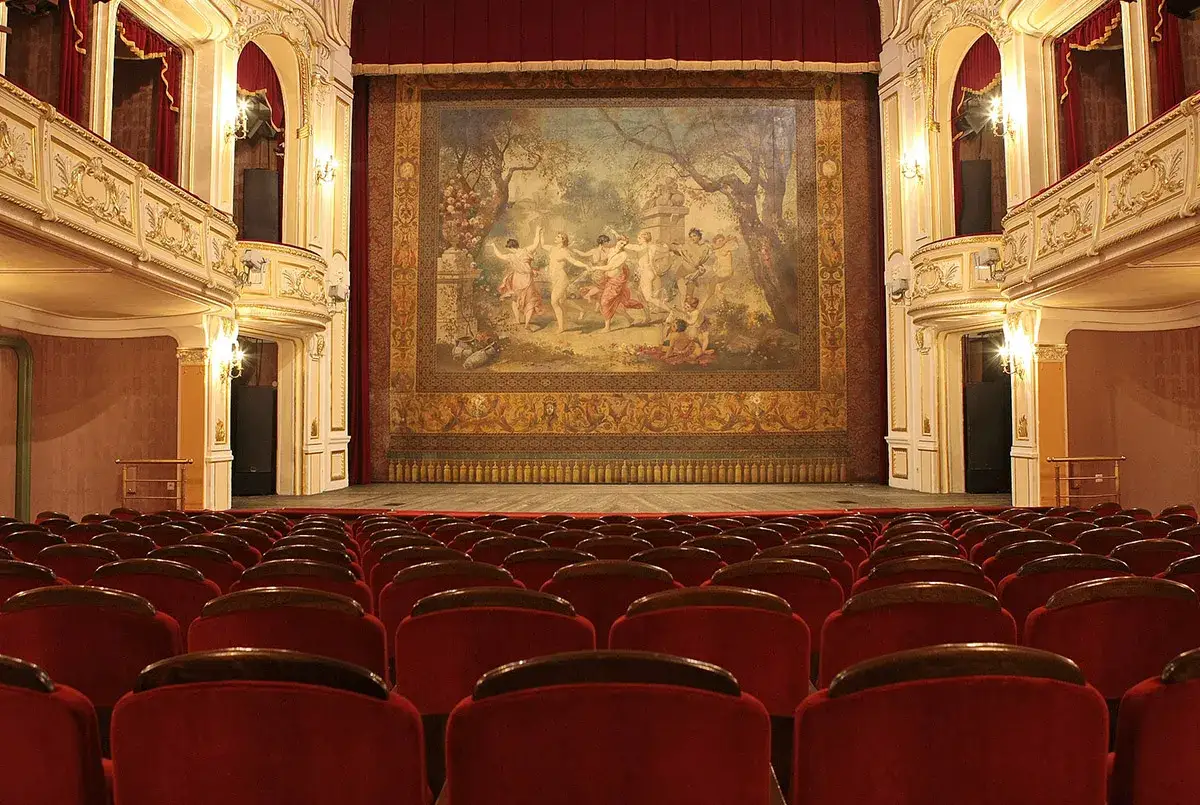 Widok na scenę i rzędy czerwonych foteli. Złocony balkon teatru ozdobiony jest malowidłem.