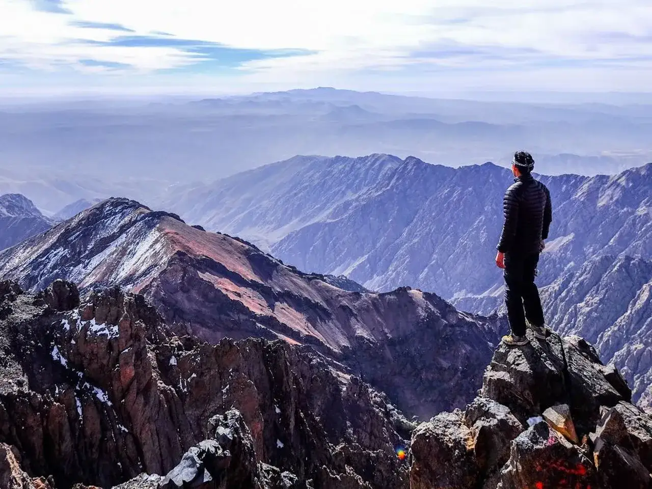 Un randonneur admire le panorama depuis le sommet du mont Toubkal, profitant de l'altitude et de la vue spectaculaire sur les montagnes de l'Atlas.