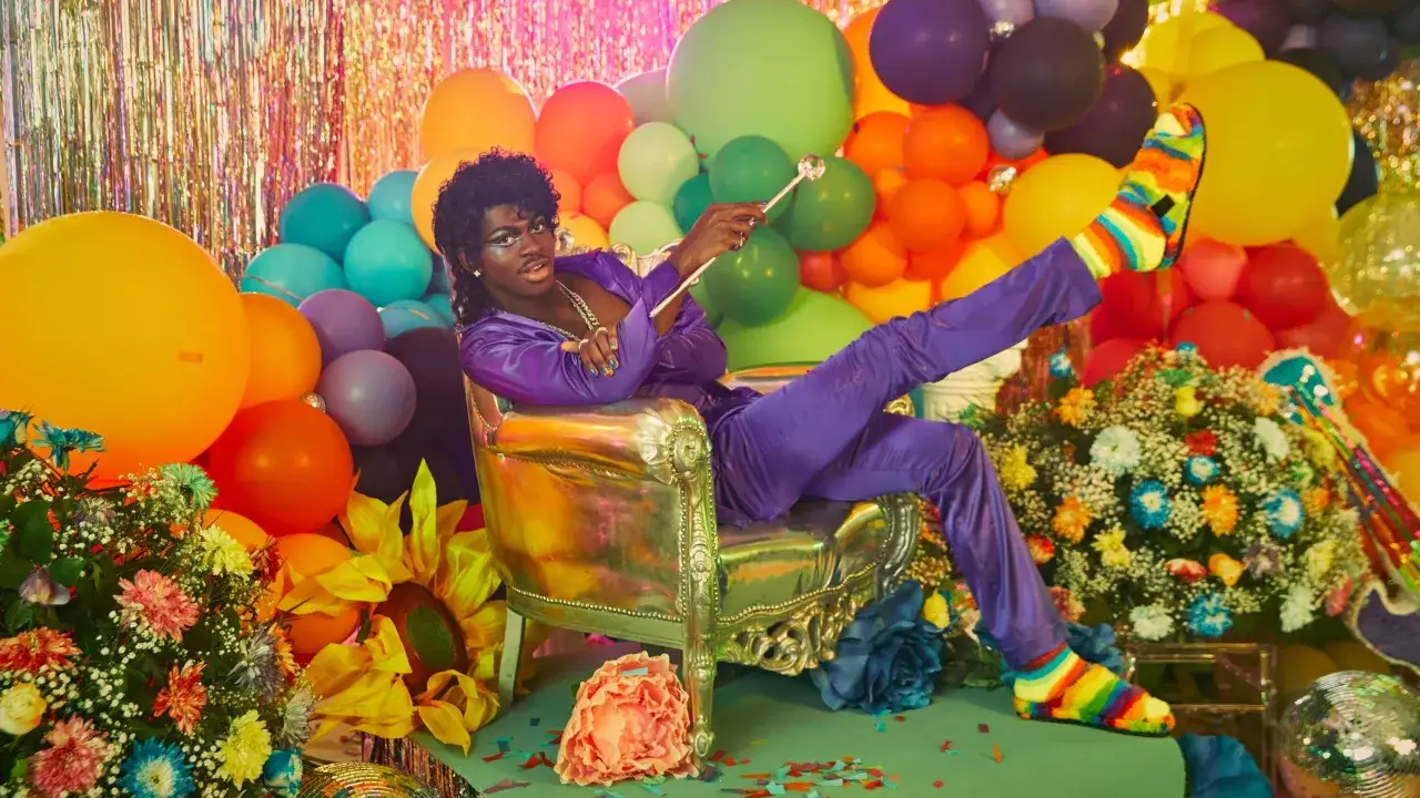 Lil Nas X w fioletowym stroju, otoczony balonami i kwiatami, emanuje pewnością siebie. Czy Lil Nas X jest gejem? Jego tęczowe skarpety i styl sugerują otwartość.